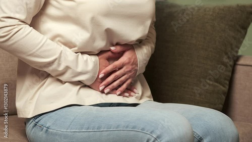 Woman experiencing stomach or abdominal pain, possibly from menstruation, indigestion, or other medical condition, sitting on a sofa and holding her belly