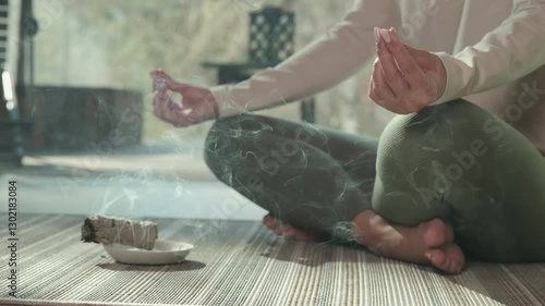 Serene woman practicing yoga in lotus position, surrounded by rising white sage smoke, embodying wellness and spiritual mindfulness indoors