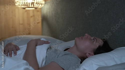 Sleepless female experiencing insomnia, tossing and turning restlessly in bed during dark night, staring blankly at ceiling with wide-open eyes while struggling with anxiety and mental exhaustion