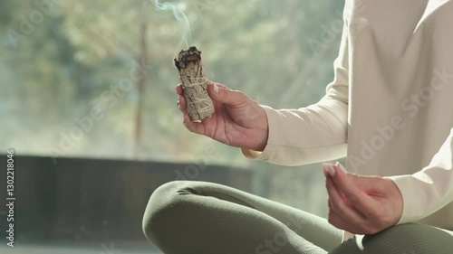 Wellness practitioner meditates in lotus position while holding a smoldering white sage bundle, using it for spiritual cleansing and energy purification
