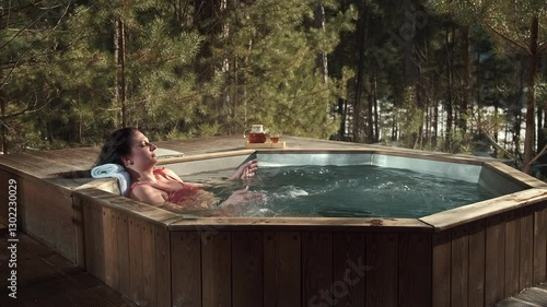 Reclining in wooden hot tub, young woman savoring tranquil moment amid snow-dusted forest landscape, sipping beverage while experiencing peaceful winter resort wellness retreat
