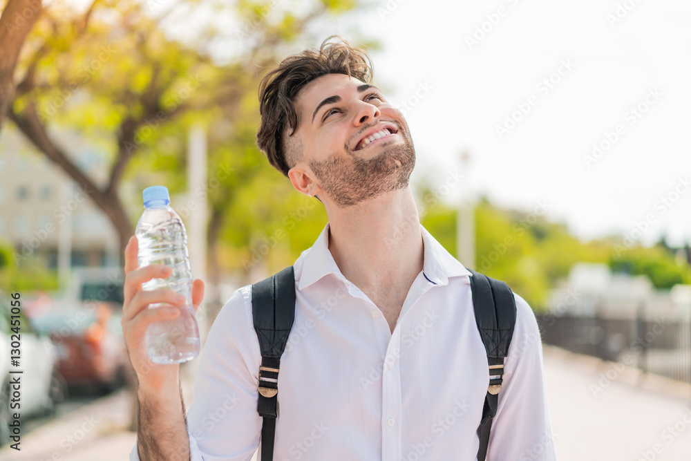Young handsome man with a bottle of water at outdoors looking up while smiling Young handsome man with a bottle of water at outdoors looking up while smiling