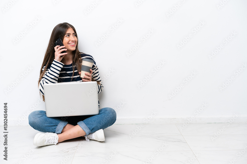 Young caucasian woman with a laptop sitting on the floor isolated on white background holding coffee to take away and a mobile Young caucasian woman with a laptop sitting on the floor isolated on white background holding coffee to take away and a mobile