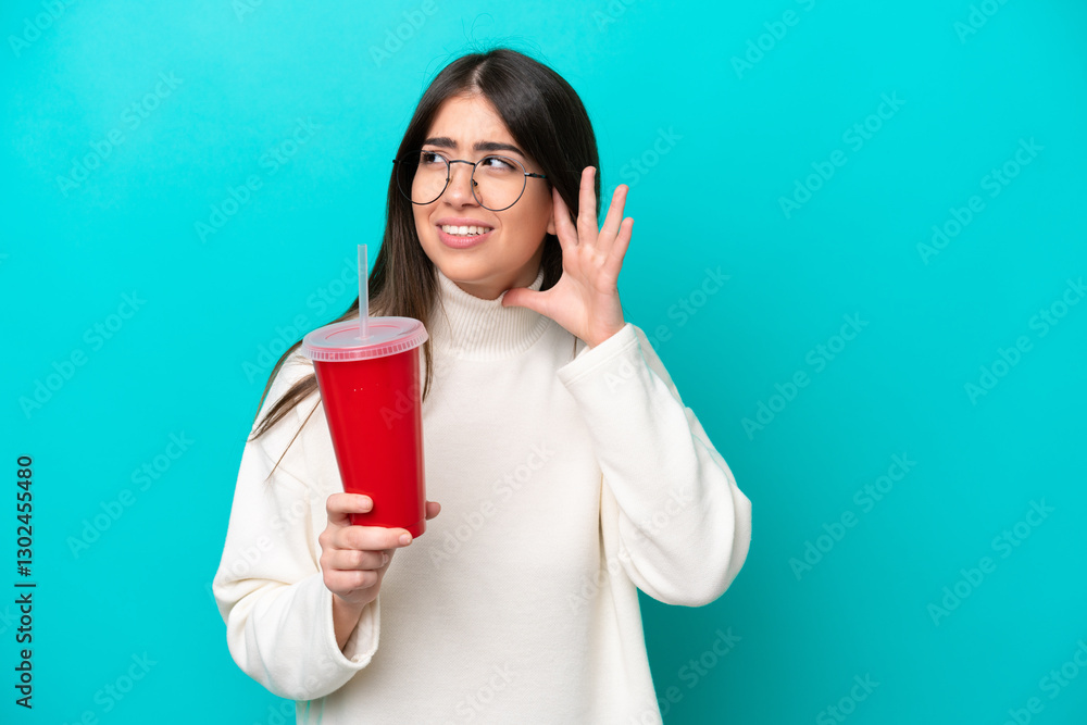 Young caucasian woman drinking soda isolated on blue background listening to something by putting hand on the ear Young caucasian woman drinking soda isolated on blue background listening to something by putting hand on the ear