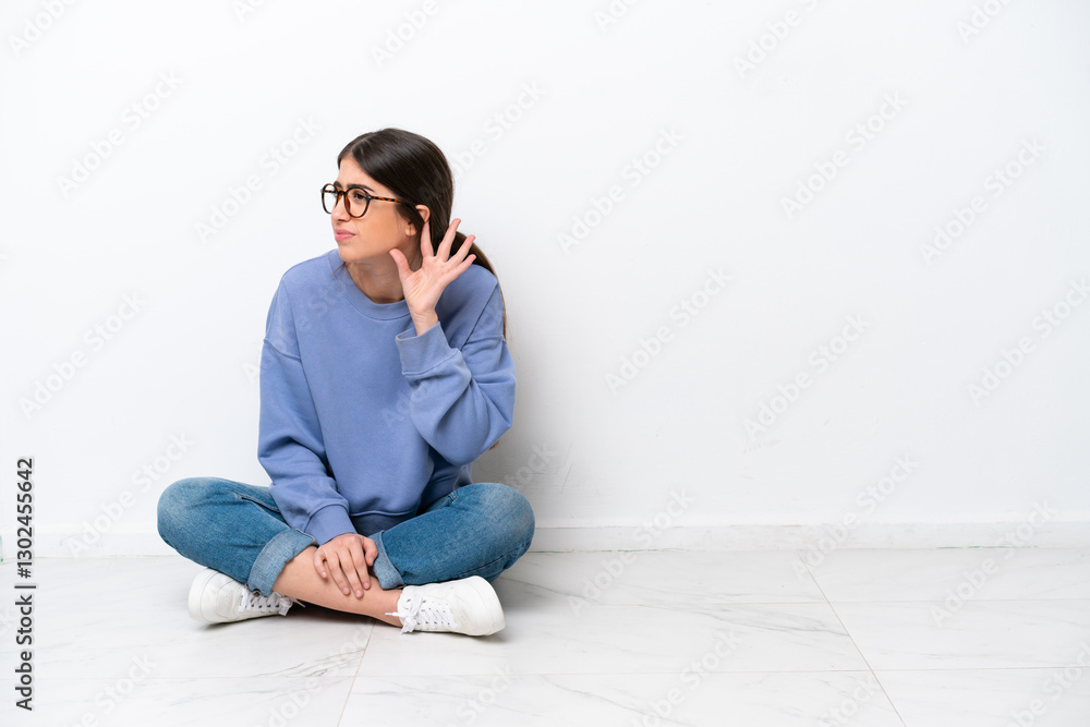 Young caucasian woman sitting on the floor isolated on white background listening to something by putting hand on the ear Young caucasian woman sitting on the floor isolated on white background listening to something by putting hand on the ear