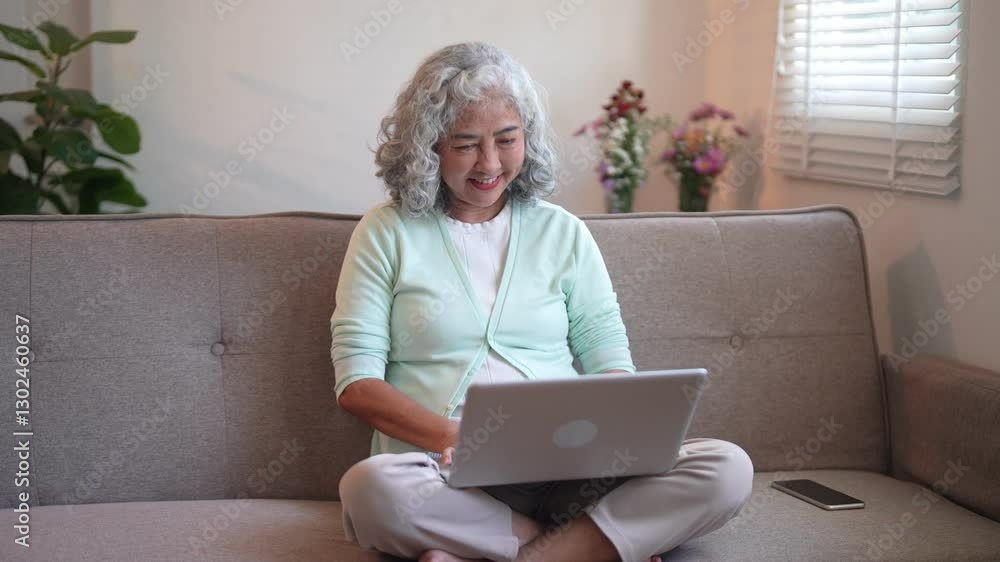 Asian senior woman using a laptop with a smile while sitting comfortably on a sofa Asian senior woman using a laptop with a smile while sitting comfortably on a sofa