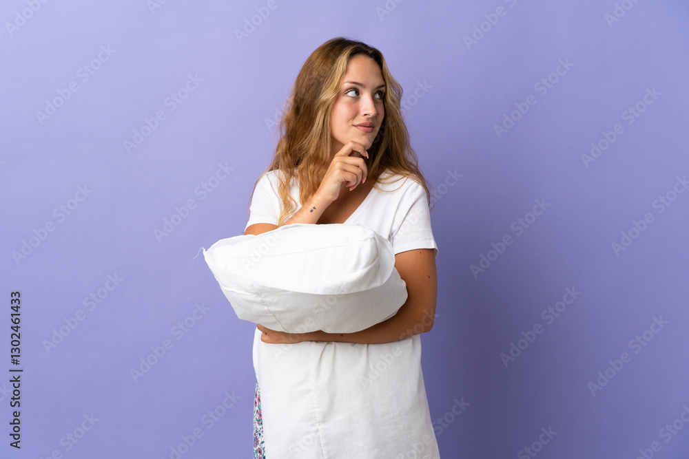 Young blonde woman isolated on purple background in pajamas and holding a pillow while thinking Young blonde woman isolated on purple background in pajamas and holding a pillow while thinking