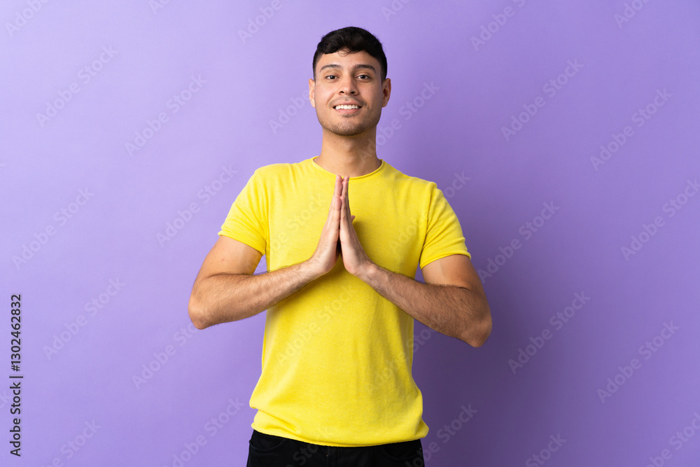 Young Colombian man isolated on purple background keeps palm together. Person asks for something Young Colombian man isolated on purple background keeps palm together. Person asks for something