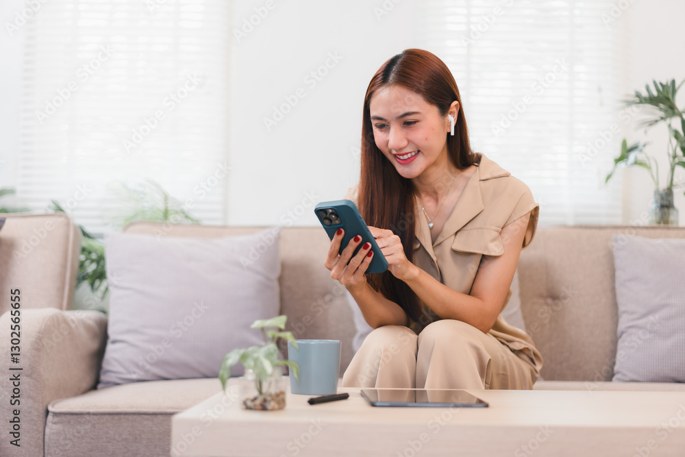 Asian businesswoman sitting on sofa, holding smartphone, appears thoughtful in cozy living room. Asian businesswoman sitting on sofa, holding smartphone, appears thoughtful in cozy living room.