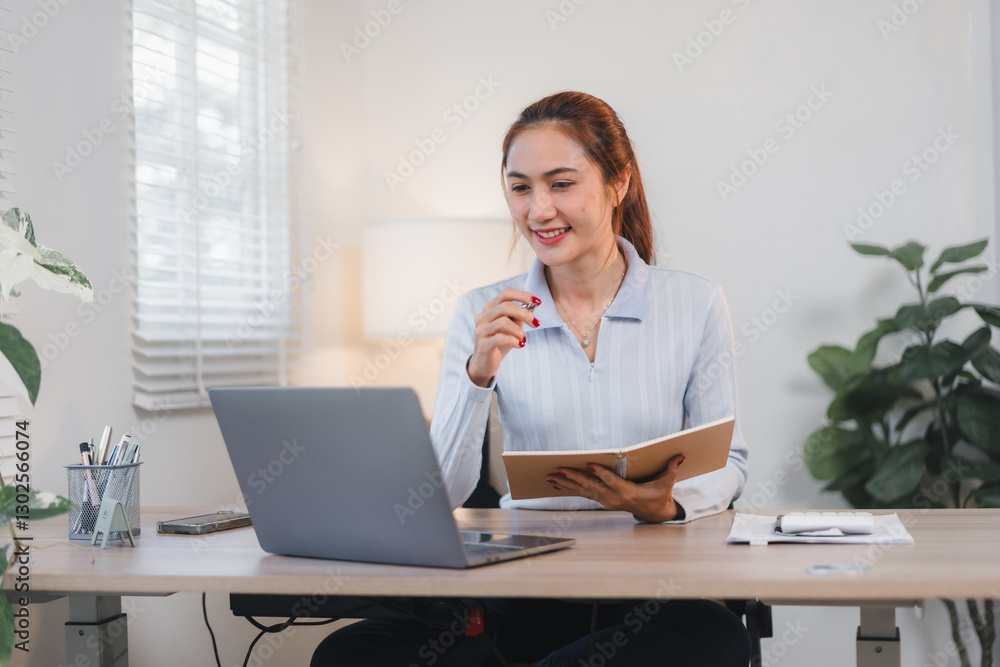 Asian businesswoman multitasking at desk, holding smartphone while writing on notepad, with laptop nearby.
