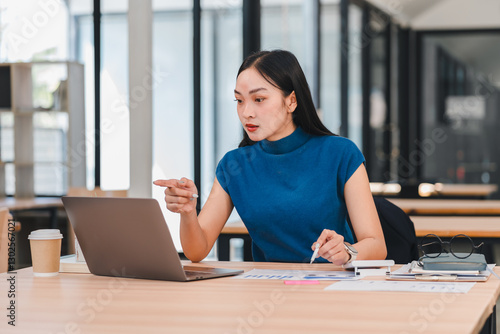Free Download Asian businesswoman in blue shirt working on laptop at desk with coffee cup and small plant. - GFXLoad