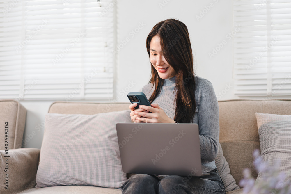 Asian businesswoman smiling while using smartphone at her desk with laptop in bright modern office. Asian businesswoman smiling while using smartphone at her desk with laptop in bright modern office.