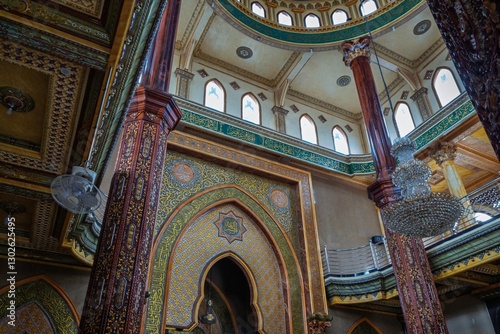Ornate Islamic architecture inside a mosque, featuring intricate arabesque patterns, Arabic calligraphy, chandeliers, and wooden columns under a domed ceiling with arched windows.