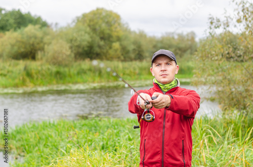 Fisherman in red sweatshirt and cap on the river bank with spinning rod in his hands.