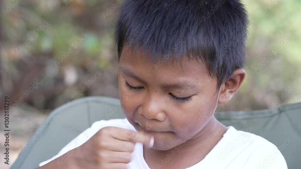 An Asian boy eats a small snack, cornflakes, savoring a candid moment of simple pleasure during a picnic or casual outdoor gathering. An Asian boy eats a small snack, cornflakes, savoring a candid moment of simple pleasure during a picnic or casual outdoor gathering.