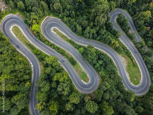 zenithal aerial view of a winding road in a green forest as cars pass by on the road in Romania.