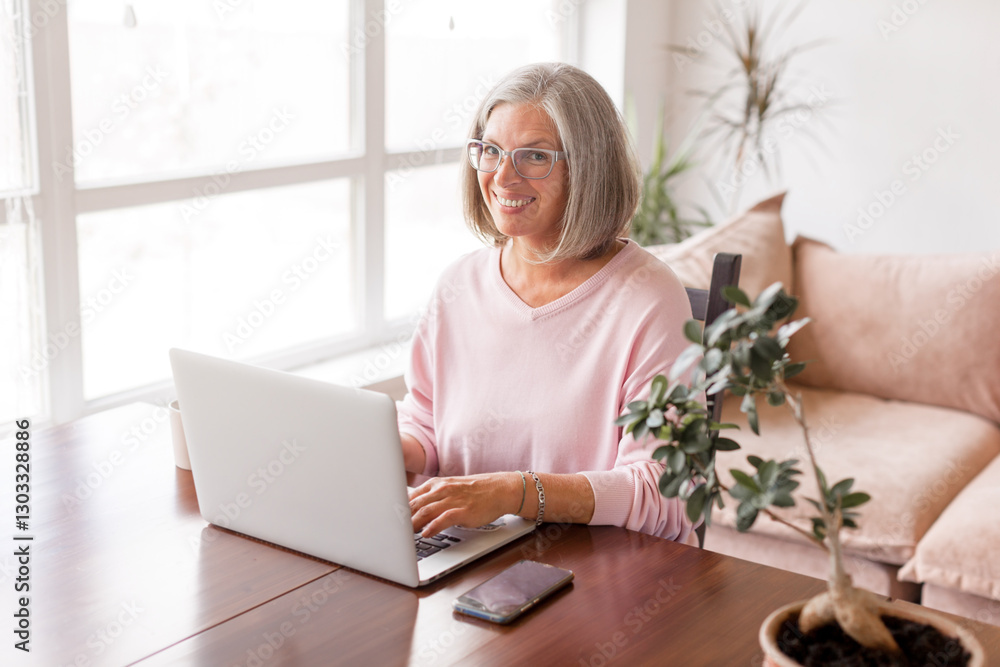 Happy middle age senior woman sitting at the table at home working using computer laptop Happy middle age senior woman sitting at the table at home working using computer laptop