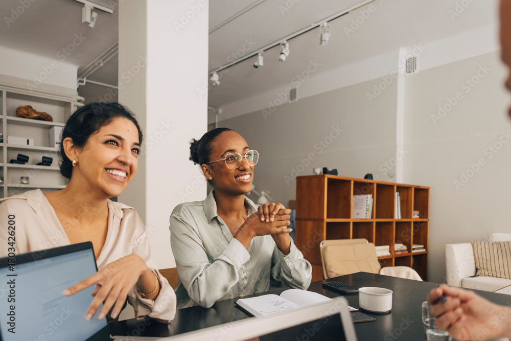 © Maskot - Happy female entrepreneurs sitting at table and discussing during business meeting in office