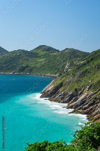 View of Guriri cove turquoise waters surrounded by Pontal do Atalaia mountains fully covered by dense green vegetation under bright summer morning blue sky in Arraial do Cabo, Rio de Janeiro - Brazil.