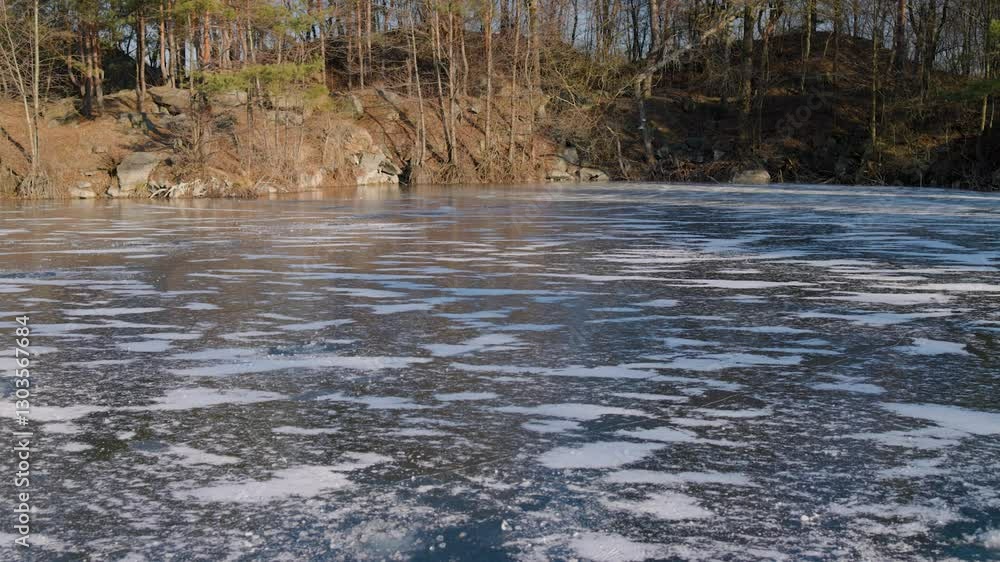 A frozen lake with a textured icy surface reflects the clear blue sky. Surrounding it, tall evergreen and leafless trees stand against a rocky shoreline, creating a serene winter landscape. A frozen lake with a textured icy surface reflects the clear blue sky. Surrounding it, tall evergreen and leafless trees stand against a rocky shoreline, creating a serene winter landscape.