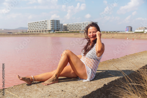 A mature woman in a white crocheted top sits on a stone ledge by a pink salt lake, basking in sunlight with her feet sandy and a serene resort landscape in the background. photo