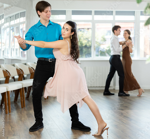 Smiling young brunette in light flowing dress performing elegant tango with stylish male partner in bright modern dance studio during group class photo