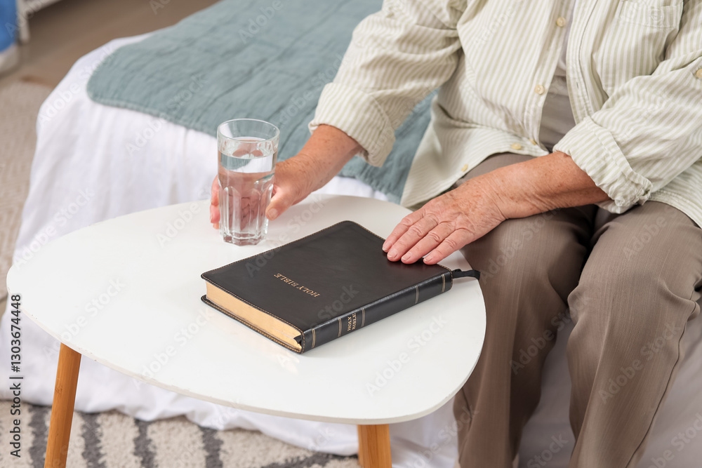 Obraz premium Senior woman with Bible and glass of water on table at home