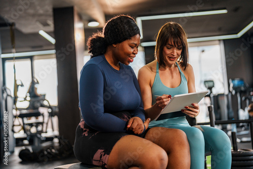 Smiling personal trainer presenting a training plan on a digital tablet to a cheerful overweight woman sitting in a gym, fostering motivation and support for her fitness journey