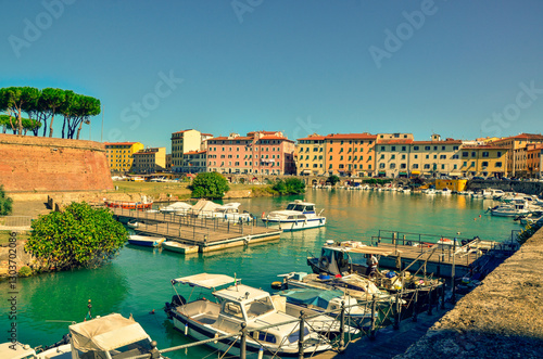 A picturesque view of a European harbor filled with boats. Colorful buildings and a historic fortress line the vibrant turquoise water under a clear blue sky.