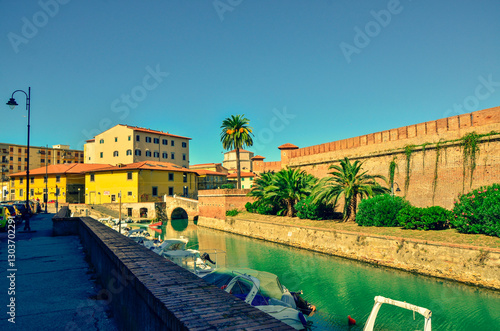 A scenic view of a city canal on a sunny day. The waterway is lined by a historic fortress wall with palm trees on one side and a bustling city quay with moored boats on the other.