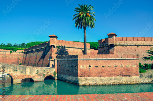 A stunning panoramic view of a historic red brick fortress, featuring a tall palm tree and a stone bridge over a turquoise moat, all under a bright blue sky.