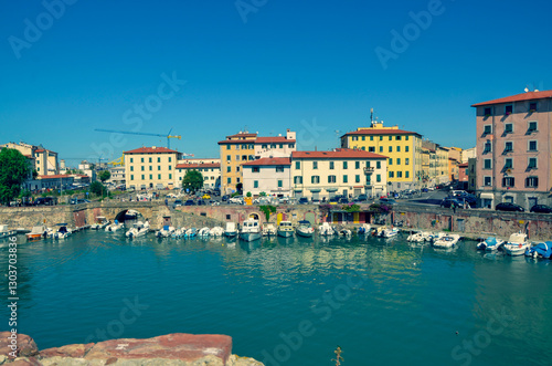 A picturesque view of a sun-drenched harbor in a historic European city. Colorful buildings line the turquoise waterway where numerous small boats are moored.