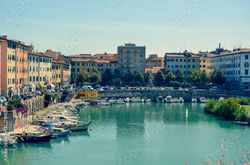 A scenic view of a wide canal in a historic European city, lined with classic buildings and numerous moored boats on a sunny day.