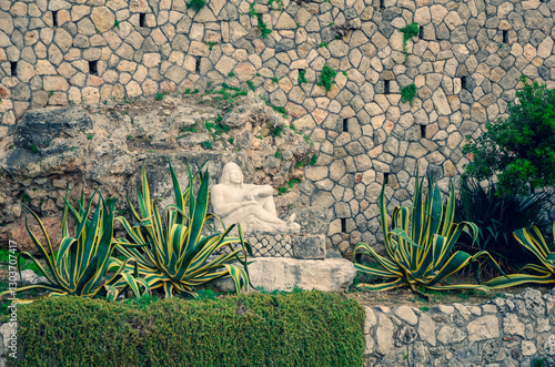 Striking variegated agave plants line a garden terrace in front of a tall, rustic stone retaining wall. A weathered white statue is nestled amongst the foliage, creating a classic Mediterranean scene.
