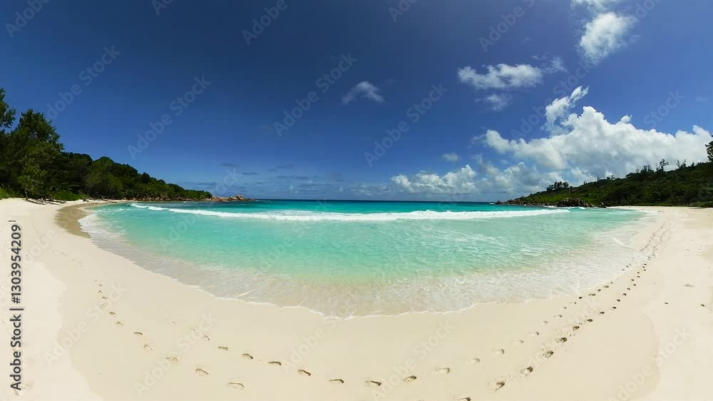 Turquoise waves rolling onto a white sand beach with footprints under a bright blue sky with scattered clouds. Anse Cocos. Seychelles, La Digue. Turquoise waves rolling onto a white sand beach with footprints under a bright blue sky with scattered clouds. Anse Cocos. Seychelles, La Digue.