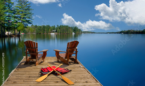 Two Adirondack chairs sit on a wooden dock overlooking the blue waters of a lake in Muskoka, Ontario, Canada. Canoe paddles and life jackets rest nearby. A cottage peeks through the lush green trees.