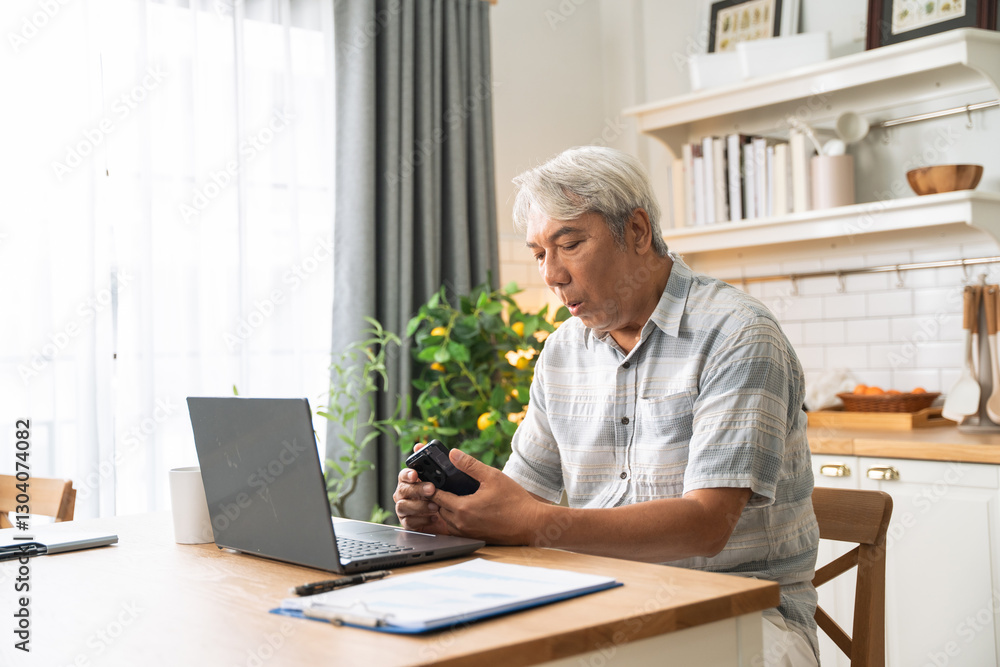 Asian senior man checking on mobile phone while working with laptop and document at home. Retired grandfather managing financial payment after retirement Asian senior man checking on mobile phone while working with laptop and document at home. Retired grandfather managing financial payment after retirement