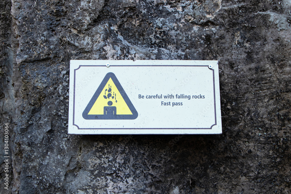 Sign warning Be careful with falling rocks fast pass to cave wall. White plastic sign with black letters and rock falls on person's head. People to strictly follow symbol. Safety of life and property. Sign warning Be careful with falling rocks fast pass to cave wall. White plastic sign with black letters and rock falls on person's head. People to strictly follow symbol. Safety of life and property.