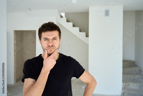 Thoughtful man in black t-shirt standing in new unfinished apartment, planning home renovation. Male home owner thinking about interior design after buying house