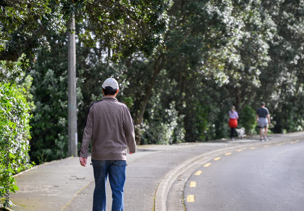 Man walking on the pedestrian sidewalk. Unrecognizable people walking the dog ahead. Auckland.