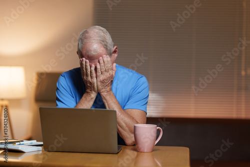 Senior man feeling stressed and exhausted while working from home, covering his face with his hands in front of a laptop, surrounded by the pressures of remote work and daily responsibilities