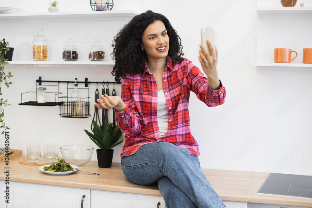 African young woman in casual outfit video chatting with cheerful expression sitting in bright kitchen. African young woman in casual outfit video chatting with cheerful expression sitting in bright kitchen.