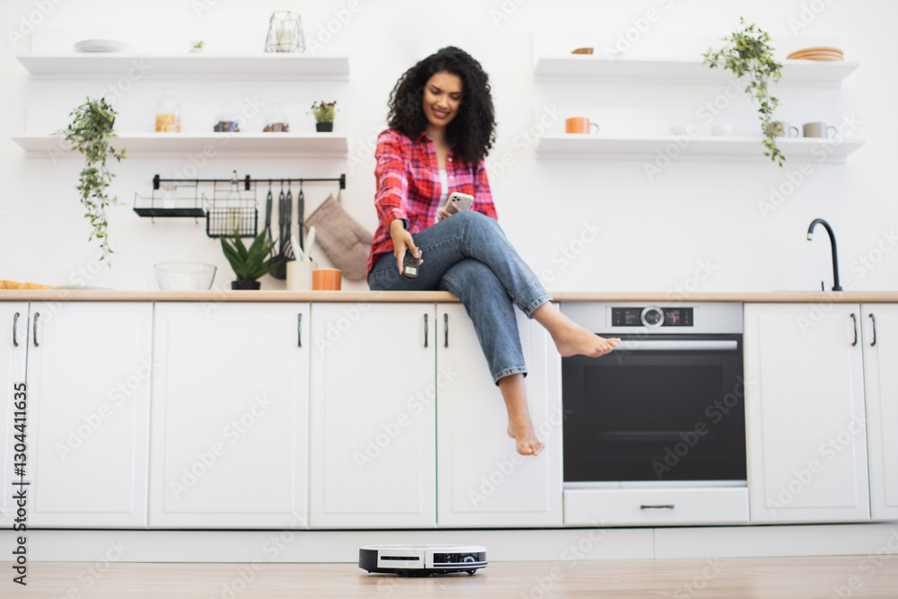 African woman sitting on kitchen counter operating robotic vacuum cleaner, looking happy and relaxed. Bright, clean kitchen environment with modern appliances showcases convenience. African woman sitting on kitchen counter operating robotic vacuum cleaner, looking happy and relaxed. Bright, clean kitchen environment with modern appliances showcases convenience.