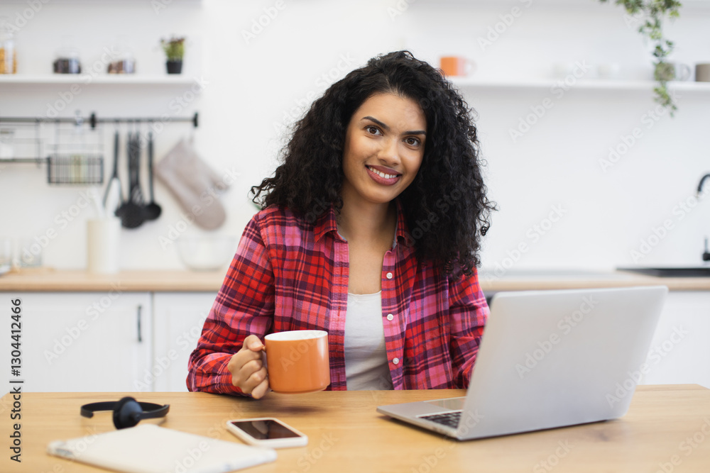 Smiling African woman with curly hair, wearing red shirt, working on laptop, holding coffee mug at home kitchen. Scene conveys comfort, productivity, and warmth. Smiling African woman with curly hair, wearing red shirt, working on laptop, holding coffee mug at home kitchen. Scene conveys comfort, productivity, and warmth.