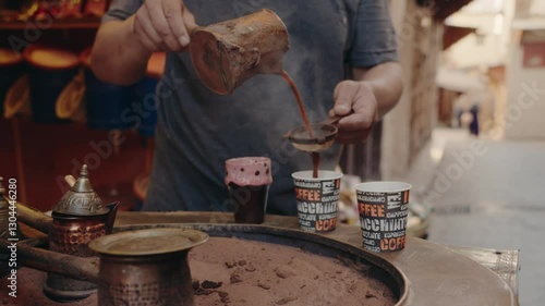 A man pours traditional Moroccan coffee through a strainer into two paper cups. He uses a vintage copper coffee pot and the cups rest on a sand-filled heater. Moroccan market.