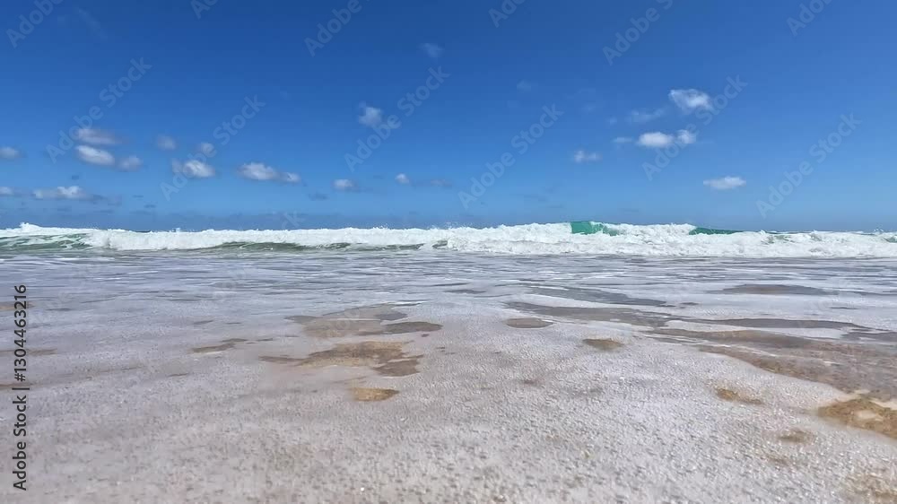 Foamy ocean waves roll onto a golden sandy beach under a bright blue sky with scattered clouds. The clear water and gentle surf create a peaceful and relaxing coastal scene. Foamy ocean waves roll onto a golden sandy beach under a bright blue sky with scattered clouds. The clear water and gentle surf create a peaceful and relaxing coastal scene.