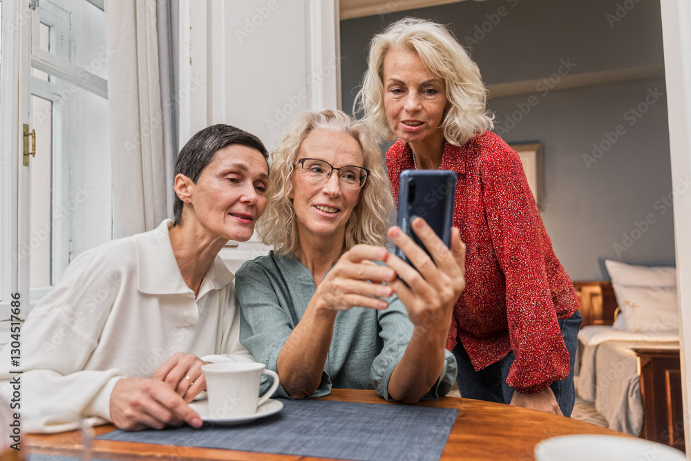 Elderly women sit at table chatting joyfully, looking in mobile telephone. Elegance blonde senior women chill together talking. Female aged friends have tea time at home and communicate smiling Elderly women sit at table chatting joyfully, looking in mobile telephone. Elegance blonde senior women chill together talking. Female aged friends have tea time at home and communicate smiling