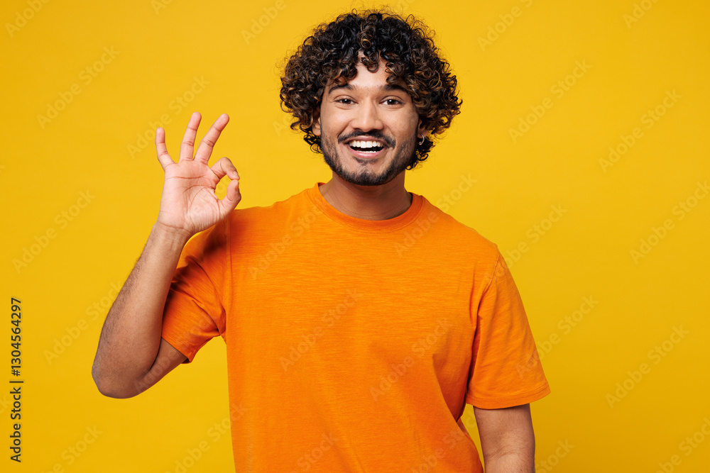 Young smiling happy satisfied cool fun Indian man he wearing orange t-shirt casual clothes showing okay ok gesture look camera isolated on plain yellow background studio portrait. Lifestyle concept. Young smiling happy satisfied cool fun Indian man he wearing orange t-shirt casual clothes showing okay ok gesture look camera isolated on plain yellow background studio portrait. Lifestyle concept.
