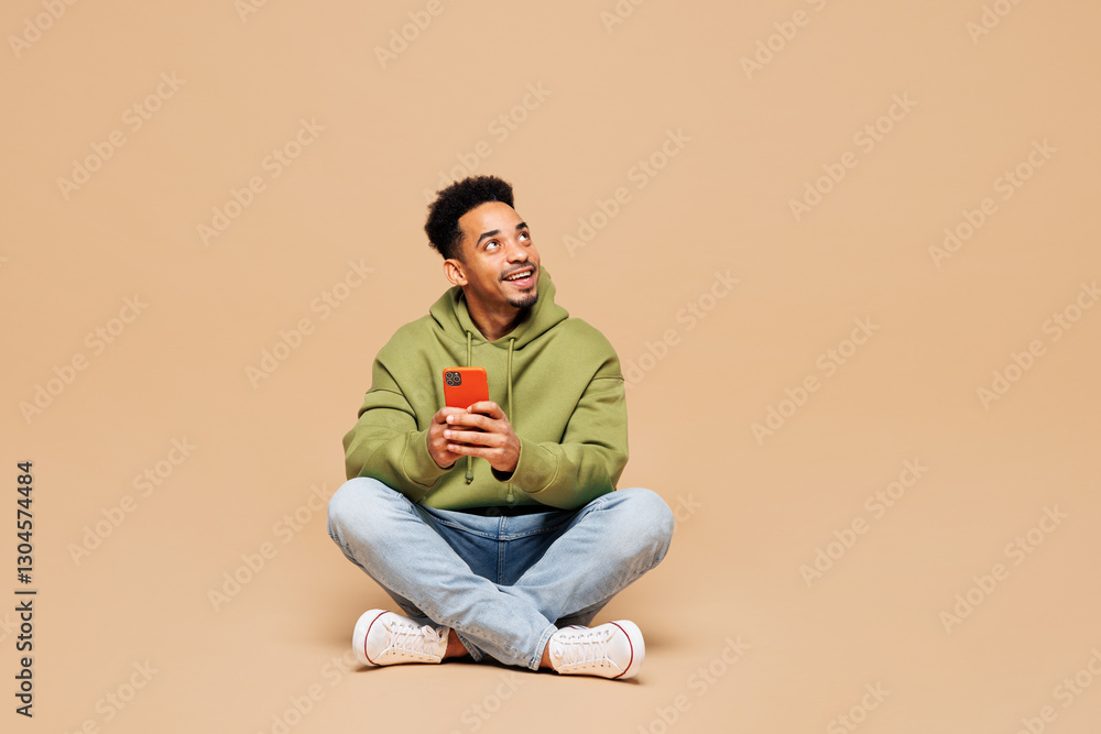 © ViDi Studio - Full body young man of African American ethnicity wears green hoodie casual clothes sit hold in hand use mobile cell phone look aside isolated on plain pastel light beige background Lifestyle concept