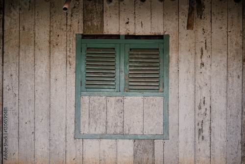 An old wooden window with faded green shutters on an old wooden house. The details of the weathered wood texture create a rustic and nostalgic rural atmosphere.